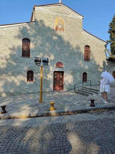 The image depicts the exterior of a stone-built church, likely in Litochoro, Greece. The architecture features a tiled roof, arched windows with intricate metal grates, and a prominent mosaic above the entrance. The facade is constructed from light-colored, rectangular stone blocks. A red door with ornate detailing is visible at the center of the building. Two individuals, dressed in white and holding a white lace umbrella, stand near the entrance, facing away from the camera. One person is holding a smartphone.
In the foreground, the ground is paved with large, irregular flagstones transitioning to cobblestones. A yellow lamppost with two spotlights stands prominently on the flagstone area. Several bollards are placed along the curb. Fallen autumn leaves are scattered on the ground and along the curb, suggesting the season. The scene is bathed in bright sunlight, casting distinct shadows from trees onto the church facade, indicating it is likely daytime, possibly late morning or early afternoon. The clear blue sky suggests fair weather. The overall atmosphere appears peaceful and serene. No visible text is legible in the image.