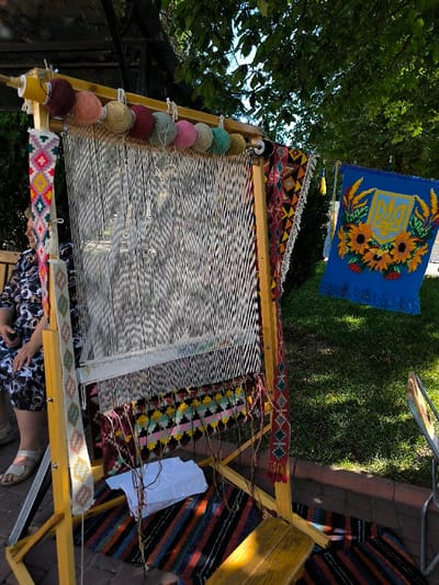 The image depicts an outdoor weaving exhibit or demonstration in Illintsi, Ukraine, illuminated by bright daylight. Central to the scene is a tall, light-brown wooden weaving loom with numerous white warp threads stretched vertically. Nine colorful skeins of yarn, ranging from maroon to black, are suspended from the top crossbar of the loom. A section of a partially woven textile, displaying horizontal geometric patterns in black, pink, yellow, blue, red, and green, is visible near the base of the warp threads. Two narrow, vertically oriented textiles with traditional geometric patterns are attached to the side supports of the loom.
To the right of the loom, a rectangular blue textile banner is suspended, featuring a central yellow Ukrainian Trident emblem surrounded by a wreath of yellow sunflowers and red berries, with a fringed bottom edge. Below the loom, a striped rug with horizontal bands of black, red, blue, and green is laid on a light-colored paved surface. White paper-like objects are situated on this rug beneath the loom's weaving area. A small, light-colored wooden stool is positioned on the striped rug to the lower right.
A person is partially visible on the far left, seated on a bench behind the loom. They wear a dark patterned dress and light-colored sandals. The background consists of green grass, bordered by a low stone or brick wall, and lush green tree foliage in the upper and upper-right portions of the frame. A dark overhead structure with a translucent roof is visible at the top, providing partial shade to the setup.