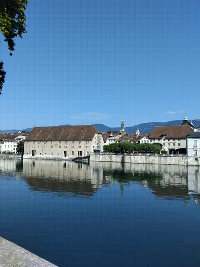 The image shows a view of the river Aare in Solothurn, Switzerland, with several historical buildings lining its bank. On the left, a large, light-colored building with a red-tiled roof dominates the foreground. This building appears to be a historic warehouse or granary, with numerous windows and shutters, and a coat of arms visible on its facade. To its right, a row of more modern buildings can be seen, including a building with the text "STADTTHEATER" visible, indicating it is a theater. A tall, slender green spire of a church or cathedral rises above the other structures in the background, along with other architectural elements of the city. The sky is clear and blue, and the river's surface reflects the buildings and the sky. A stone embankment is visible in the lower part of the image. The overall scene suggests a sunny day in a picturesque European city.