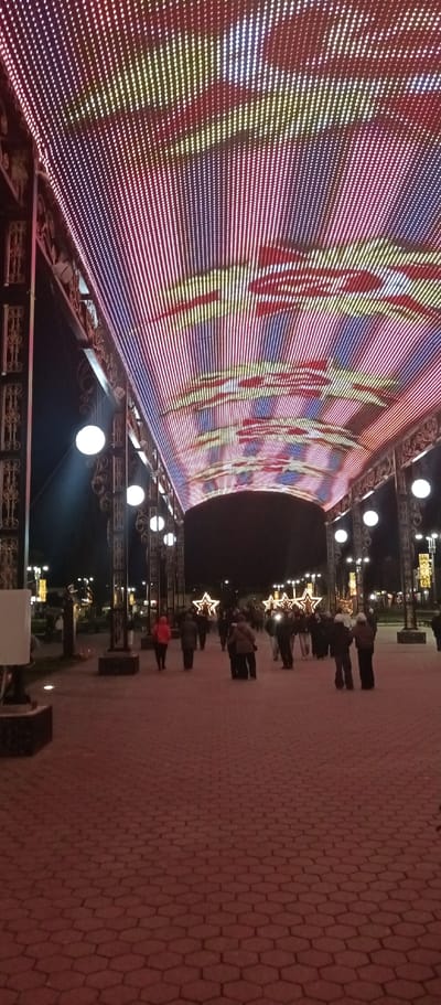 The image captures a nighttime scene in Irbit, Russia. A pathway paved with red, hexagonal bricks stretches into the distance, where a crowd of people is gathered. Overhead, a canopy of closely spaced LED lights creates a tunnel of vibrant colors including red, yellow, and blue, in a striped pattern. The canopy is supported by ornate, dark metal structures, each adorned with spherical white lamps. Further down the pathway, lighted star decorations add to the festive atmosphere.