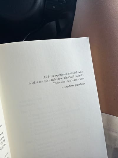 The image shows a page from a book open to a quote by Charlotte Joko Beck: "All I can experience and work with is what my life is right now. That's all I can do. The rest is the dream of ego." The book is angled slightly, resting near what appears to be a car interior, including a steering wheel with buttons and a portion of the dashboard. A person's arm and part of their clothing (white) are also visible at the top of the frame. The context provided mentions Toseong-myeon, South Korea, but no element visible in the image confirms this location.