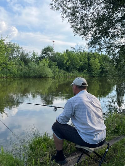 A man is fishing on the bank of a calm body of water, with lush green trees and vegetation lining the opposite bank. The sky above is partly cloudy, with a distant radio tower visible on the horizon. The man is seated on a portable folding stool, facing away from the camera, towards the water. He is wearing a striped long-sleeved shirt, dark shorts, a white baseball cap, and black socks with shoes. Two fishing rods are visible, one extending into the water. The water reflects the trees and sky. The location is identified as Суроватиха, Russia.