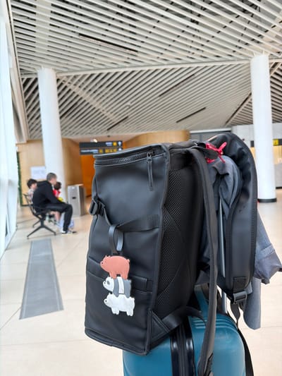 The image depicts a black backpack with a "We Bare Bears" cartoon keychain attached, placed on top of a blue suitcase in an airport terminal. The background shows a high-ceilinged terminal space with a repeating angled white facade, a floor grille, and blurred figures sitting in the distance, partially obscured. An airport sign with directions is visible on the terminal wall.