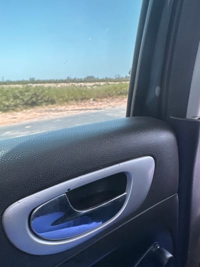 The image depicts a view from the interior of a vehicle in Dokwa, Nigeria, looking out through the side window. The foreground features the upper portion of a car door panel, primarily textured black plastic or faux leather. A chrome-plated interior door handle, set within a silver trim bezel, is centrally located in the lower half of the frame; a blue reflection is prominent on its surface, and minor scratches are visible on the trim. Outside the vehicle, a paved road stretches horizontally across the midground, bordered by a dirt shoulder. Beyond the road, the background consists of an open, vegetated landscape under a clear, bright blue sky, indicating an outdoor, daylight setting. No individuals or specific actions are discernible.