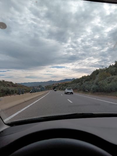 A car travels on a highway under a cloudy sky. The view is from the driver's perspective, featuring the road, adjacent trees, and other vehicles in the distance. A section of the car's steering wheel is visible on the right, while the dashboard runs along the bottom. The sky is predominantly covered with dense clouds.