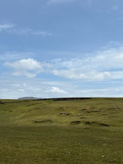 The image depicts an open, natural landscape in Bluff Cove, Falkland Islands (Islas Malvinas). The upper two-thirds of the frame consist of a light blue sky with scattered white cumulus clouds, denser near the horizon. The lower third of the frame shows an undulating terrain covered in green grass. A distinct, dark, irregular horizontal line marks the distant horizon, appearing as a raised edge or cliff. A dark, rounded landmass or distant hill is visible on the far left, partially obscured by atmospheric conditions. Within the grassy foreground, several small, irregular patches of reddish-brown earth or rock are exposed. No individuals, animals, structures, or actions are present in the scene.