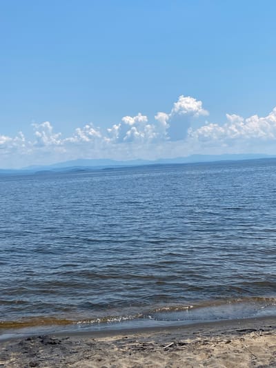 The image shows a tranquil scene of a body of water, likely a lake or ocean, bordering a sandy shore. The water is a deep blue, with gentle ripples creating a textured surface. In the distance, a hazy silhouette of land can be seen, suggesting a vast expanse of water. The sky above is a clear, bright blue, dotted with fluffy white clouds that form a striking contrast against the azure backdrop. The clouds are clustered along the horizon, adding depth and visual interest to the sky. The shoreline on the right side of the image is composed of light-colored sand, with some scattered debris, hinting at a natural, undeveloped setting. The overall impression is one of peace and serenity, with the natural elements of sky, water, and sand harmoniously coexisting. The lighting suggests it is daytime, likely mid-morning or late afternoon, given the soft quality of the light. The location is identified as the Town of Peru, United States of America. There is no text visible in the image.