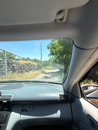 The image is taken from the inside of a vehicle, looking forward through the windshield. The dashboard is visible in the foreground, a gray plastic surface with air vents and control buttons. The windshield shows a rural scene. A dirt road leads away from the vehicle, bordered by a dry, grassy area and a low, rough stone wall on the left. A metal fence runs along the wall. Beyond the wall, there are trees and vegetation under a clear blue sky. A utility pole is visible on the right side of the road. The exterior shot captures a serene, undeveloped area, likely a countryside road. There are no people visible in the primary scene. The location is described as being in the city of Kosh, Armenia, suggesting this is a view of a road within or on the outskirts of this Armenian city. The timestamp on the image indicates it was captured on 2023-07-07.