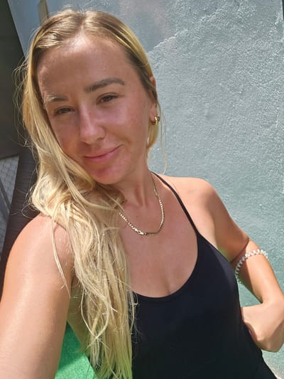 A young woman with blonde hair is taking a selfie outdoors, standing against a light blue wall. She is wearing a black tank top and a gold necklace. On her left arm, she wears a white bracelet. Sunlight illuminates her face and hair, casting shadows on the wall. The image appears to have been taken during the daytime. The location is not specified in the image.