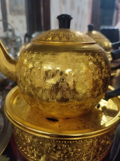 This image showcases an ornate, gold-colored teapot with intricate floral patterns. The teapot has a black lid and handle. It rests on a matching gold-colored circular tray, which is itself placed on a decorative gold-colored bowl. The background is blurred, suggesting an indoor setting, possibly a display or collection of decorative items. The lighting is somewhat dramatic, with highlights reflecting off the metallic surfaces, indicating a well-lit environment. The focus is entirely on the teapot and its accompanying serving pieces, emphasizing their craftsmanship and decorative quality. There are no people visible, and the scene appears to be a static display rather than an active event. The detailed etching on the teapot and bowl suggests a traditional or antique aesthetic. No visible text is present in the image. The location context provided is Blang Cut, Indonesia, which might imply the origin or style of these items, perhaps reflecting local artistry or cultural heritage.