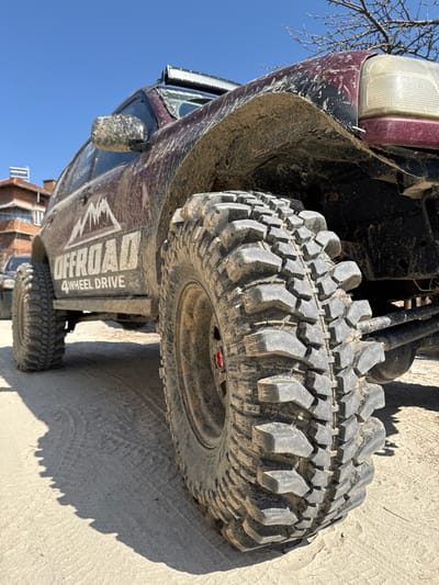 A heavily modified, mud-splattered, dark red or maroon SUV is parked on a light-colored dirt or sandy surface under a clear blue sky in Breznitsa, Bulgaria. The vehicle features large, aggressive mud-terrain tires on black wheels, with a red center cap visible on the front wheel. A white decal on its side reads "OFFROAD 4 WHEEL DRIVE" with a mountain graphic. A prominent LED light bar is mounted on the roof above the windshield. Extensive mud and dirt are caked onto the vehicle's body, fenders, tires, and side mirrors, indicating recent off-road activity. The front passenger-side headlight assembly is visible. Long shadows are cast on the ground from the vehicle and its tires, suggesting direct sunlight. In the background to the left, a portion of a brick building with terracotta roof tiles is visible, along with another partially obscured vehicle. Bare tree branches are visible against the sky in the upper right. No individuals are present.