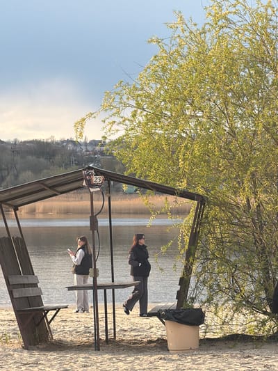 The scene is captured on a sunny day near a body of water, possibly a lake or a wide river, in Olginskaya, Russia. The image includes a man and a woman standing at the edge of the water, with a structure resembling a pier or a gazebo partially visible in the foreground. The gazebo has a sign with a raccoon illustration and the number "23". The terrain appears to be a sandy beach. The surrounding environment includes trees with budding leaves and a view of buildings on a distant hillside.