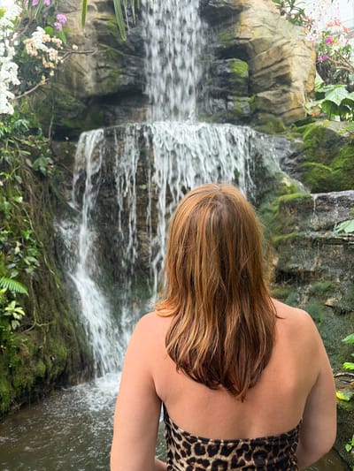 The image depicts a woman with auburn hair, seen from the back, wearing a leopard print strapless bikini. She is standing in front of a multi-tiered waterfall, surrounded by lush greenery and moss-covered rocks. The waterfall cascades down a rocky cliff face adorned with various flowering plants, including white and pink orchids. The water collects in a pool at the bottom, where the woman is positioned. The scene is set in an indoor or sheltered environment that replicates a naturalistic tropical setting. This location is identified as the Queen Sirikit Botanic Garden in Mae Rim, Chiang Mai province, Thailand.
