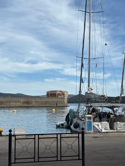 This landscape-oriented image captures a tranquil harbor scene in Portoferraio, Italy, under a bright blue sky dotted with scattered white clouds. In the foreground, a black metal railing with decorative square patterns runs along a concrete pier, where a blue and white utility box (likely for power/water) is also visible.

Several sailboats are moored alongside the pier, their tall masts and intricate rigging extending towards the sky. On the stern of one prominent sailboat, the name "CHIA" is clearly visible in blue script, along with a partial registration number "17G...". This boat, like others, is equipped with black fenders along its hull. Another sailboat has a grey cover over its boom, and small colorful pennants flutter from the rigging.

The calm, light blue water of the harbor reflects the sky and the boats. Several yellow cylindrical buoys float on the water's surface. Dominating the midground is a robust, historic stone building, likely a fortress or tower, characterized by its reddish-brown stone and arched openings. This structure sits at the end of the pier or on a breakwater. In the background, green, tree-covered hills rise gently. The scene depicts a peaceful daytime setting, with no visible human activity, on a clear and pleasant day.