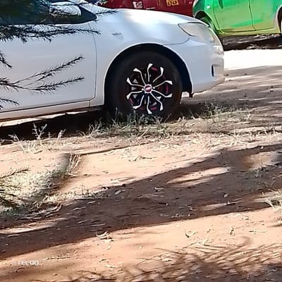 The image displays the front left quarter of a white sedan, featuring a black, branch-like decal along its visible side. The front wheel is equipped with a black tire and a decorative wheel cover with a multi-spoked design in red, white, and black. The vehicle is positioned on an uneven dirt surface sparsely covered with dry grass. Long, dark shadows extend across the foreground from left to right. In the background, the rear section of a red vehicle with a partially visible license plate "KDC 624P" is present, behind which a portion of a green vehicle can also be seen. No people are visible. This scene is located in Machakos Central ward, Kenya.