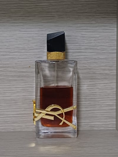 A horizontal, rectangular glass perfume bottle with a black cap and gold textured band lies on a wood-grain surface. The bottle is partially filled with a dark amber liquid. A prominent gold YSL logo is affixed to the front of the bottle. The logo appears partially submerged in the liquid. The background consists of light gray wooden paneling with subtle vertical lines.