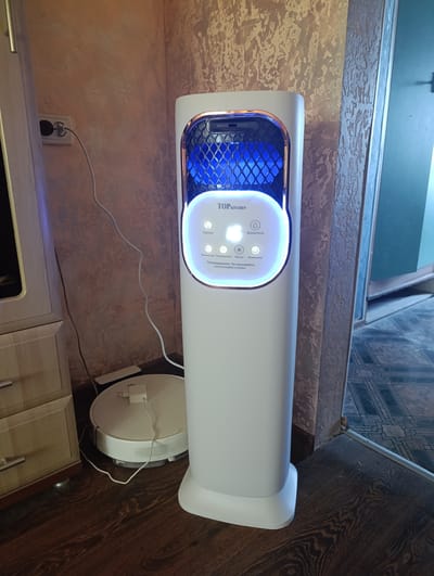 The image shows an indoor scene in Ladyzhyn, Ukraine. The main subject is a tall, white, modern-looking air purifier with a blue, diamond-patterned light panel at the top. The purifier has a display screen with illuminated text, including "TOP KITCHEN" and some Russian words that appear to be labels for functions like "Светло" (Light) and "Влажность" (Humidity). Below the screen are icons, one of which resembles a snowflake.
To the left of the air purifier is a wooden cabinet with drawers and metallic handles. A white power cord snakes from a wall outlet, plugged into a white charging base for a round, white robotic vacuum cleaner positioned on the dark wooden floor.
The wall behind the air purifier and cabinet has a textured wallpaper with a pattern of abstract, overlapping shapes in shades of beige and brown. To the right of the air purifier, a doorway is visible, leading to another room with dark patterned flooring and a green door. The lighting in the scene suggests it is daytime, likely indoors. There are no people visible in the image. The overall impression is of a domestic setting, possibly a living room or hallway, with functional appliances present.
