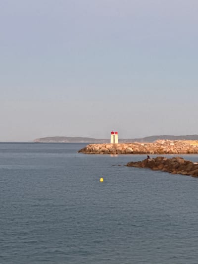 This image shows a view of the sea and a breakwater with a lighthouse in Le Lavandou, France. The breakwater is made of large rocks and has a small lighthouse with red and white stripes at its top. There are two people sitting on the rocks near the lighthouse. A yellow buoy floats in the water to the left of the breakwater. In the distance, there is a hazy coastline with hills. The sky is clear and light blue.