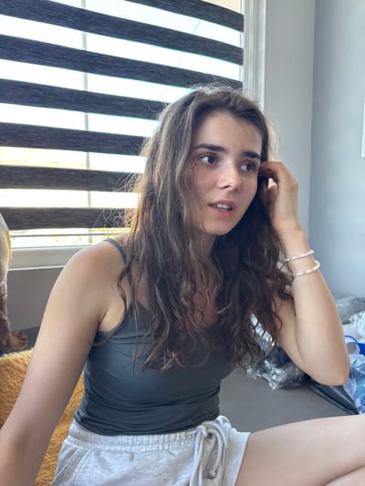 The image shows a young woman with long, wavy brown hair, looking to her right with a surprised expression. She is wearing a dark gray tank top and light gray sweat shorts. She has a bracelet made of small white beads on her left wrist. She is seated on a couch, with a yellow blanket visible to her left. Behind her, there are horizontal blinds on a window. To her right, there are some objects wrapped in plastic and a blue plastic object, possibly a container. The setting appears to be indoors, likely a residential room. There is no indication of the specific city of Dibich, Bulgaria in the image.