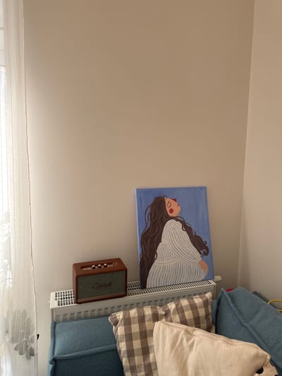 A painting of a woman laying down with long brown hair on a blue background is hanging on a beige wall. Below the painting is a brown Marshall speaker mounted on a white radiator. A bed with blue and patterned pillows is situated next to the radiator. A white curtain is visible at the bottom of the image. There is no evidence to indicate this scene is in Люк, Russia.