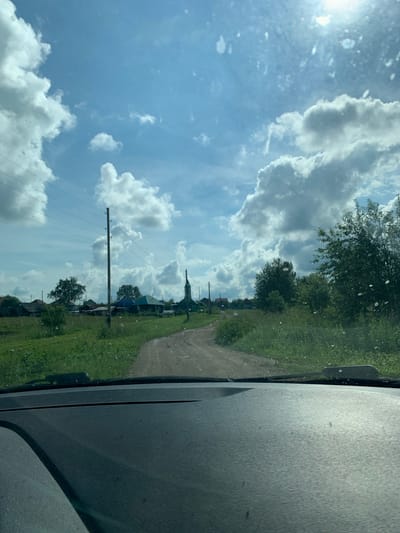 The image captures a view from inside a vehicle, looking out through a dusty windshield. A dirt road winds through a rural landscape with green grass and trees on either side. In the distance, a small village is visible with several houses and what appears to be a church with a steeple. Telephone poles and power lines run along the left side of the road. The sky is blue with large, fluffy white clouds. The overall impression is of a sunny day in a quiet, rural area, possibly in the city of Barsai, Russia, given the context.