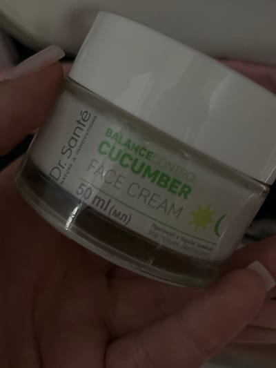 A close-up, angled shot shows a jar of Dr. Santé Balance Control Cucumber Face Cream held in a person's hand. The jar is mostly white with green lettering. The text "Dr. Santé NATURE & INNOVATIONS" is visible on the left side, and "BALANCE CONTROL CUCUMBER FACE CREAM" is on the front. Below that, "50 ml (мл)" is written. A yellow starburst and a green crescent shape are also visible on the jar. The background is blurred, showing hints of fabric and what might be furniture, suggesting an indoor setting. The hand holding the jar has long, light-colored fingernails, and the skin tone suggests a female. No specific details about the city of Pănășești, Moldova are visible in this image.