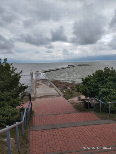 A long pier extends into the choppy, grey waters of the Curonian Lagoon under a heavily overcast sky. Red brick stairs and walkways lead down to a concrete path that merges with the pier. Dense green trees frame the left and right sides of the scene. The water is disturbed by wind, creating waves that crash against the shore and the pier. Several individuals are visible on the pier, appearing to be standing or walking. The timestamp "2026-07-05 18:34" is superimposed on the lower right corner of the image. The location appears to be the Ventė Cape, a prominent point on the Lithuanian coast known for its bird observatory and lighthouse, which would explain the pier extending into the lagoon.