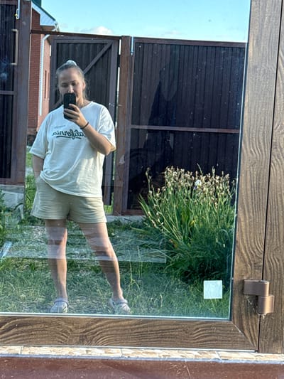The image is a reflection in a window of a young person holding a phone and taking a selfie. The subject is wearing a light-colored t-shirt with some text on it and light-colored shorts. They have their hair pulled back. Their reflection shows them standing with one leg extended, appearing to be in mid-stride. The reflection also shows a background of green grass and a dark wooden fence. In the foreground of the reflection, there are some dried flower stalks. The window is framed by a dark, textured material that resembles wood, with a metal hinge visible at the top. The overall scene suggests an outdoor setting during the daytime, with natural light. The reflection is slightly distorted by the glass. The text on the t-shirt appears to be a stylized script, possibly a brand name or phrase, but it is not entirely legible. The location is indicated as Pychanki, Russia.