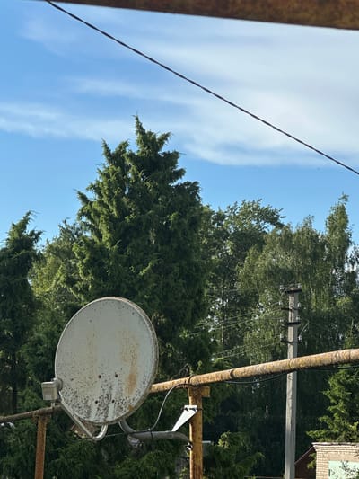 The image depicts an outdoor scene in Gavrilov Posad, Russia, under a bright blue sky with thin white clouds. In the foreground, a rusty metal pipe runs horizontally across the frame, supporting a weathered, off-white satellite dish on the left side. The satellite dish is visibly stained and corroded. Behind the dish and pipe, a dense line of various green trees dominates the midground, with a prominent tall evergreen tree on the left and other deciduous trees to the right. A concrete utility pole with insulators and wires is visible behind the trees on the right. A thin black wire stretches diagonally across the upper portion of the image. The lower right corner shows a section of a brick building with a green element.