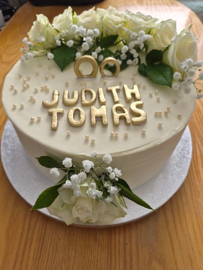 A white, round cake with a smooth, off-white frosting is presented on a white cake stand. The cake is adorned with gold lettering that spells out "JUDITH TOMAS". Two gold rings are placed above the names. The cake is decorated with white roses and baby's breath flowers, with green leaves peeking out from beneath. The cake is situated on a wooden surface, possibly a table, with a visible wood grain. The background is blurred, but appears to be an indoor setting. There are no people visible in the image. The scene is static and appears to be a celebration or special occasion cake. No specific location in Šilutė, Lithuania can be determined from the image.