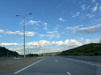 The image captures a view of a highway in Otarsheni, Georgia, under a bright blue sky dotted with fluffy white clouds. The highway stretches into the distance, with clear lane markings indicating multiple lanes of travel. On either side of the highway, there are concrete barriers and green, tree-covered hills. Several lampposts line the side of the road. In the background, wind turbines are visible on the horizon, suggesting a rural or semi-rural setting. The scene appears to be captured from a moving vehicle, as indicated by the slight blur and the perspective.