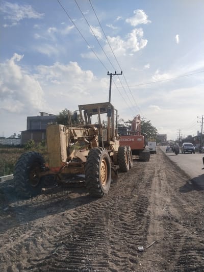 The image depicts a construction scene in Barokulamgajah, Indonesia. A yellow Komatsu grader and an orange Hitachi excavator are parked on a dirt patch alongside an asphalt road. Tire tracks are visible in the dirt. Utility poles and power lines run overhead. A few vehicles, including a car, a truck and motorcycles, are on the paved road, with pedestrians. In the background, there is a building and vegetation. The sky is partly cloudy.
