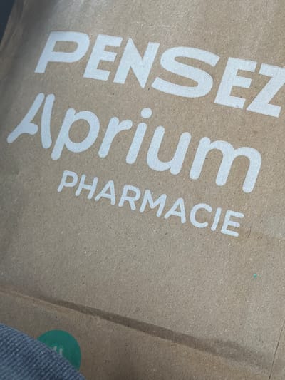 The image shows a close-up of a brown paper bag with white lettering. The words "Pensez Aprium Pharmacie" are printed on the bag. The bag is angled, and part of a blue-grey fabric is visible in the lower left corner. The background is out of focus. The image does not contain any information about the city of Ödsmål, Sweden.