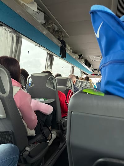 This media file captures the interior of a passenger bus, likely a long-distance coach, showing passengers seated and traveling. The image is taken from a slightly elevated, rotated perspective, looking down the aisle towards the front of the bus.
In the immediate foreground, a person's blue jacket, featuring a prominent white Nike swoosh logo, hangs or is worn by someone out of frame. The bus interior consists of rows of grey, upholstered seats with plastic armrests and back shells. Several passengers are visible, mostly from the back or side. One person in the middle distance wears a red and black striped top. Further down the aisle, heads of other passengers are visible, some with grey hair, suggesting a mix of age groups. One passenger is seen with a pink neck pillow or plush toy resting on their shoulder, suggesting they are resting or sleeping.
Overhead luggage racks, featuring a light grey base with a striking blue fabric strip, line the sides of the bus. Various bags, clothing items, and a white shopping bag with a black spot pattern are stowed in these racks. The bus windows are covered with light-colored curtains, and the outside view shows an overcast or bright, cloudy sky, indicating daytime. Air vents are visible along the ceiling. Below one seat in the foreground, a white sneaker and a black bag are discernible. A small device, possibly a power outlet or entertainment screen, with a red button and some indistinct text, is attached to the back of a seat.
The scene depicts people undertaking a journey by bus, likely in Rogozinovo, Bulgaria, as per the provided context. The general atmosphere appears calm, with passengers settled into their seats for travel.