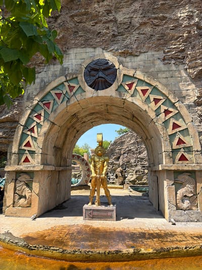 The image shows a golden statue of a warrior positioned under an ancient-looking stone archway. The archway is decorated with triangular patterns in red and green, and features a carved, dark face above the opening. The statue appears to be made of metal and is holding a weapon. Flanking the archway are stone carvings of mythical sea creatures. Beyond the arch, a rocky, natural landscape is visible, with some greenery and a small body of water in the foreground. This scene is located within the Movieland Park in Castelnuovo del Garda, Italy, specifically in the area known as the "Hollywood Premiere" zone.
