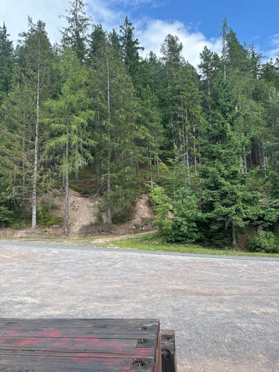 The image depicts a section of a forest with a gravel parking area in the foreground. The forest is dominated by tall evergreen trees with dense green foliage. The ground slopes upwards into the forest, showing exposed soil and some undergrowth. The sky above is blue with scattered white clouds. In the immediate foreground, the edge of a wooden structure, possibly a bench or a platform, is visible. The structure is made of dark, weathered wood with some red markings. The overall scene is natural and appears to be an outdoor recreational area. This location is consistent with the natural landscape surrounding Chełmsko Śląskie, Poland, known for its forests and hilly terrain.