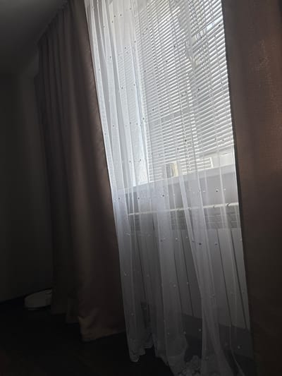 The image shows a window with white horizontal blinds covered by sheer white curtains adorned with small, scattered pearls. To the left of the sheer curtains are thick, dark brown curtains. In the lower left corner, partially obscured by the dark curtains, is a white robotic vacuum cleaner. The scene appears to be an interior space, possibly a living room or bedroom, in Kinel'-Cherkasy, Russia. The lighting suggests it is daytime, with sunlight filtering through the blinds. There are no people visible in the image.