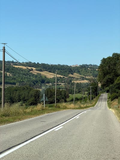 This is an eye-level, full shot of a rural road winding through a hilly landscape under a clear blue sky. The road is asphalt with double white lines marking the center and a single white line on the right edge. The road curves to the right and ascends into the distance. On the left side of the road, there are power poles and lines. Beyond the road on the left, there is a grassy verge, followed by dense green trees. The background is dominated by rolling hills covered in a patchwork of green forests, yellow fields, and some scattered buildings. The overall impression is of a sunny day in a countryside setting in Oyeu, France.