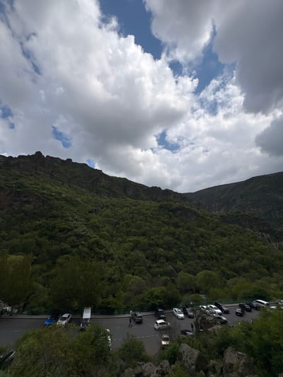 An eye-level, outdoor shot shows a vertical landscape of the city of Goght, Armenia. A mountain range dominates the left portion of the frame, its rocky face partially covered in lush, green foliage. The sky above features a mix of dark, heavy clouds and patches of blue. To the right, a road runs parallel to the mountain's edge, lined with parked cars and greenery. The road appears to be wet, suggesting recent rainfall. A few indistinct figures of people are visible on the roadside. The overall environment suggests a tourist spot or scenic overlook.