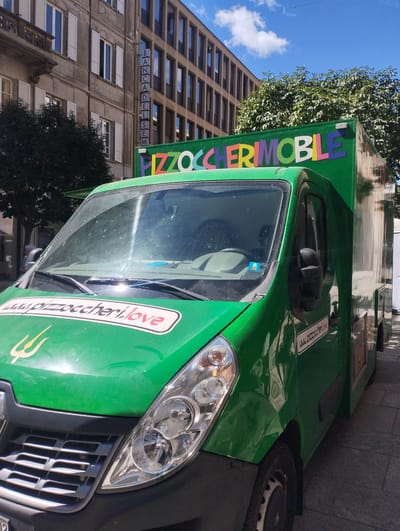 A bright green food truck is prominently featured, parked on a paved street under a clear blue sky with sparse white clouds. The truck is likely a mobile food vendor, specializing in "Pizzoccheri," a type of pasta, as indicated by its name.
On the front top edge of the truck's canopy, the word "PIZZOCCHERIMOBILE" is displayed in colorful, varied letter styles. The hood of the truck features a white oval sticker with "www.pizzoccheri.love" written in red and black, below which is a golden trident-like logo. The same website address is visible in smaller black text on the front passenger side panel.
In the background, two buildings stand out: an older, light-colored building with white window shutters on the left, and a modern, multi-story brown building with numerous windows on the right. A vertical sign on the modern building reads "BANCA DEL SEM". Green foliage from trees is visible behind and beside the truck.
The scene depicts an urban environment, likely a city street or square in Bellinzona, Switzerland, given the Italian language visible on the signs. The bright sunlight indicates daytime. The truck is stationary, suggesting it is either open for business or parked. No explicit activity is taking place, and no individuals are clearly visible.