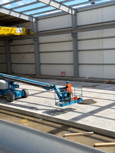 The image depicts the interior of a large, unfinished industrial building with a metal frame structure and white insulated walls. A yellow overhead crane is visible in the background, labeled with "10.0t". In the foreground, a blue boom lift, identified as "4x4", is positioned on a concrete floor. A construction worker wearing a blue helmet and an orange safety vest is standing in the basket of the boom lift, appearing to be operating it. The environment is characterized by concrete flooring, exposed metal beams, and a partially constructed roof structure. Sunlight casts shadows across the floor. In the distance, a red and white safety barrier is visible. There is some water or liquid on the concrete floor in the foreground and a plastic water bottle. The location is identified as Skrzydlna, Poland.