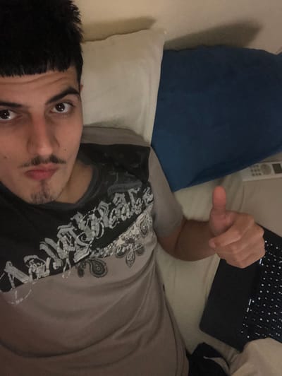 The image shows a person lying on a bed with two pillows, one blue and one white. A laptop is positioned near the person's head. The person is giving a thumbs-up. The person has dark hair, brown eyes and a moustache and is wearing a gray shirt with black designs. There is a remote control lying by the blue pillow. It is not possible to identify the location as the city of Beccar, Argentina, based on the provided image.