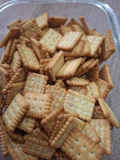 A close-up, overhead shot shows a clear plastic container filled with golden brown square crackers. The crackers are piled high, with some overlapping and others lying flat. They have a slightly textured surface and are arranged in a chaotic, yet appealing, manner. The lighting is even, highlighting the crisp edges and the subtle variations in color of the crackers. The background is blurred, drawing the viewer's attention to the main subject. There is no discernible indication of the city of Gorkovskiy, Russia, in the image.