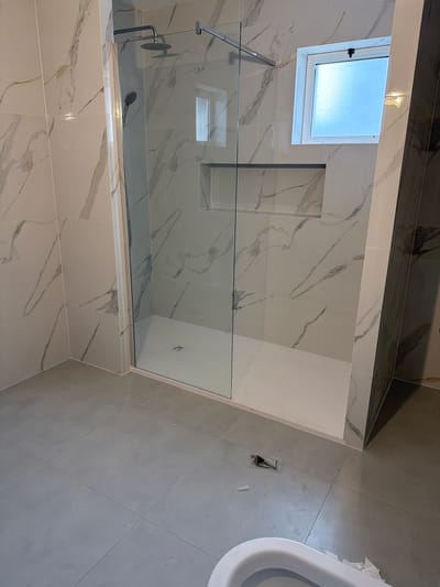 The image shows a modern bathroom with a glass-enclosed shower. The walls are tiled with large, white marble-patterned tiles with grey and gold veining. The shower floor is a white, rectangular tray with a central drain. Above the shower, there is a built-in niche shelf. A small, frosted window with a tilt mechanism is visible on the tiled wall. The floor of the bathroom is covered with large, grey square tiles. In the foreground, a portion of a white toilet bowl is visible, along with some crumpled white paper and a small clear plastic package containing dark-colored items, possibly plumbing or electrical components. The overall impression is of a newly renovated or constructed bathroom, likely in a residential property in Gondomar, Portugal, as indicated by the context.