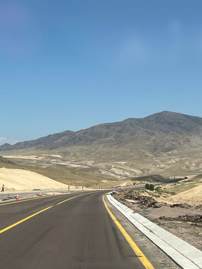 The image captures a partially constructed roadway in a desert-like mountainous landscape. The road, paved with dark asphalt and marked with yellow lines, curves to the right, leading towards distant hills. Construction cones and barriers are visible along the roadside, indicating ongoing work. In the distance, a few vehicles are seen on the road, and the terrain is characterized by dry, undulating hills and sparse vegetation. The sky is a clear, bright blue. The scene appears to be located in the vicinity of Алғабас, Kazakhstan, a region known for its vast, arid landscapes and ongoing infrastructure development.
