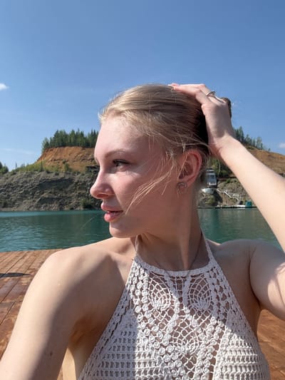 The media file shows a young woman with blonde hair, wearing a white crocheted top, outdoors by a body of water. She is leaning forward, with her right arm raised, hand resting on her head. A ring is visible on her left ring finger. She is wearing a circular earring on her visible ear. The background consists of a steep, rocky cliff covered in trees, with a calm body of water in the foreground. The sky is a clear, bright blue, suggesting a sunny day. The scene appears to be a recreational area, possibly a lake or reservoir, given the presence of boats and docks visible in the distance on the water. The overall mood is relaxed and summery. No visible text is present.