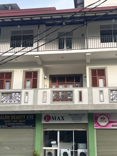 The image shows the exterior of a multi-story building with shops on the ground floor. The building has a white facade and a red tiled roof. There are several windows on the upper floors with wooden shutters. The ground floor features three storefronts. The leftmost store, labeled "SALON BEAUTY EYE," appears to be a beauty salon, offering bridal, hair, and beauty services. The middle store is an appliance repair shop, listing services for various electronics like TVs, audio systems, washing machines, fans, microwaves, ovens, toasters, blenders, and electric kettles. The rightmost storefront displays a pink logo with the name "LOSELKA." The shops are located at 691/A, Udugama Road, Walahanduwa, Galle, Sri Lanka. Two washing machines are visible through the open doorway of the middle shop. Power lines are strung across the upper portion of the building.