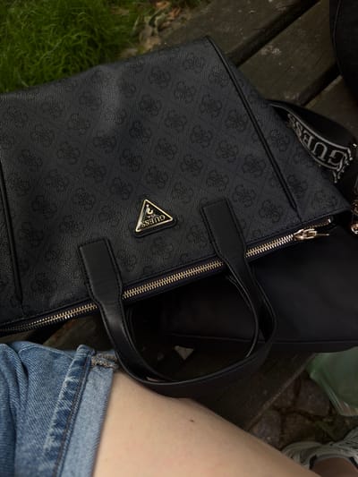 An overhead shot captures a black Guess handbag with a monogram pattern resting on a wooden bench. A person's legs, clad in denim shorts, are visible in the lower left of the frame, resting against the bench. The background shows a patch of green grass to the left of the bench. The scene appears to be outdoors, possibly in a park or public seating area. No specific location in the city of Herford, Germany is identifiable from the visual information provided.