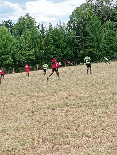 The image depicts an outdoor football match in progress at a dry, grassy field in Lwandeti ward, Kenya. Approximately 10-12 individuals, primarily young males, are visible. Players are divided into two teams, one wearing red shirts and black shorts, the other wearing green shirts and black shorts. A player in a red shirt is in the foreground, running right, appearing to approach a white football positioned centrally on the field. Another player in a green shirt is further back, also moving right. Other players are scattered across the field, engaged in various stages of play. A few spectators are visible in the far background, near the treeline, observing the game. The field's ground cover is sparse, dry, brown grass with exposed earth. A dense line of tall green trees forms the background. The sky above is partly cloudy with white and grey clouds against a light blue hue, indicating daytime conditions.