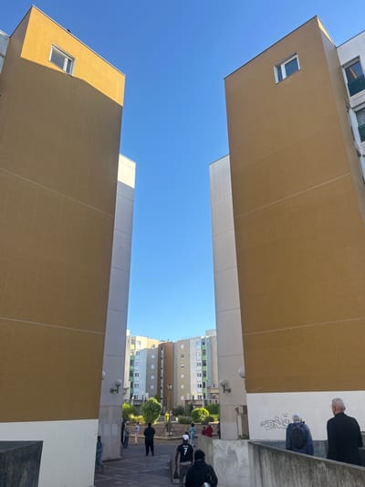 This upward-looking, portrait-oriented image captures a dense urban environment under a clear blue sky. Two prominent, multi-story buildings, characterized by their striking golden-brown tiled facades, flank the central void, with their lower sections featuring white or light grey cladding. Rectangular windows with white frames are visible on both structures.
In the mid-ground and background, further apartment buildings are visible, appearing in shades of grey, white, and some with green panels, typical of modern residential complexes. A clear, bright blue sky fills the space between the structures, indicating a sunny day.
Several people are gathered on what appears to be an elevated walkway or courtyard area to the right side of the frame. In the upper right, an older man with grey hair, dressed in a dark coat, stands alongside a person in a blue jacket and white cap leaning against a white wall. This wall features black graffiti. Further down the walkway, other individuals are present, including one person wearing a black top with "VINI JR" visible in white text on the back, and another in a red outfit. The people appear to be socializing or passing through the area.
Notable details include outdoor wall lights on the mid-ground buildings and some scattered greenery like trees and bushes. The strong sunlight casts shadows, suggesting midday or early afternoon. The architecture and density of the buildings are consistent with the provided location context of Noisy-le-Grand, France, a commune known for its residential developments.